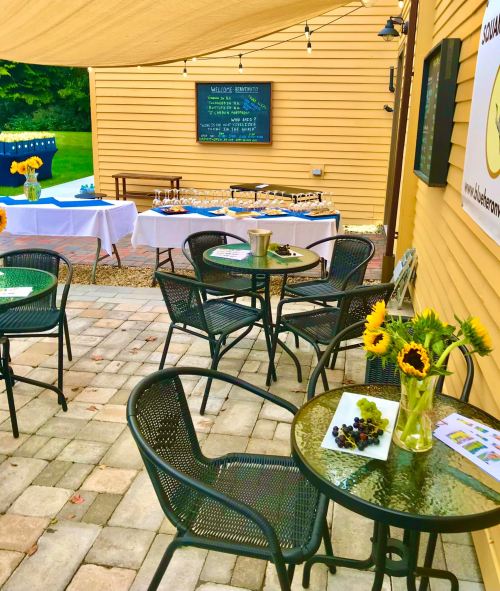 Outdoor seating area with tables, sunflowers, and a dessert display under a canopy, set against a yellow building.