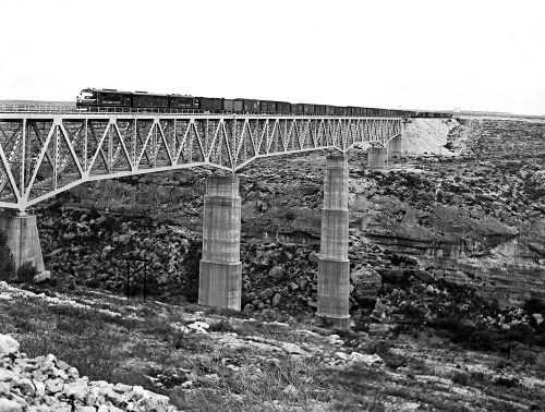 A train crosses a large metal bridge over a rocky canyon, captured in black and white.