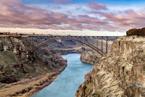 A large metal bridge spans a river, surrounded by rocky cliffs and a cloudy sky.