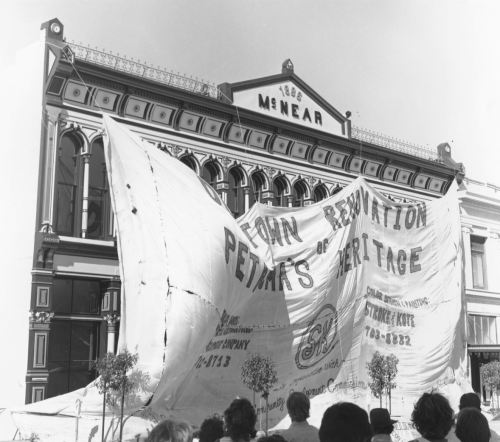 A large banner reading "Restoration of Heritage" covers a historic building, with a crowd gathered in front.