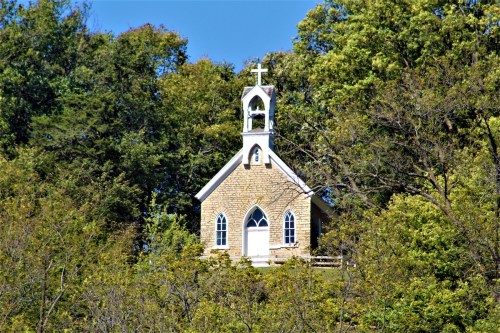 A small stone church with a bell tower, surrounded by lush green trees under a clear blue sky.