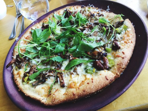 A freshly baked pizza topped with greens, mushrooms, and cheese on a round plate, set on a wooden table.