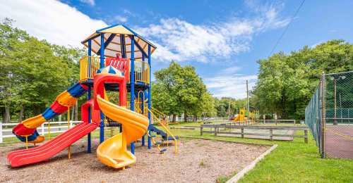 Colorful playground with slides and climbing structures, surrounded by green trees and a fenced area in the background.
