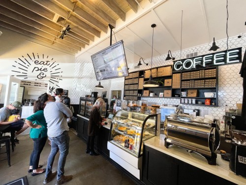 A cozy café interior with a coffee counter, pastries on display, and customers waiting in line. Bright, inviting atmosphere.
