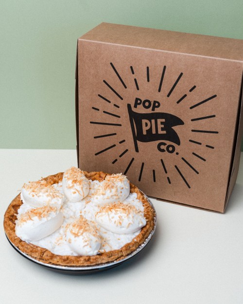 A coconut cream pie with whipped topping sits next to a brown box labeled "Pop Pie Co." on a light background.