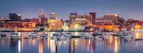 City skyline at dusk with illuminated buildings and boats docked in a calm harbor.