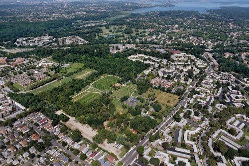Aerial view of a green park surrounded by residential areas and cityscape, with a river visible in the distance.