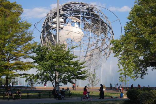 A large metallic globe sculpture surrounded by trees and people enjoying a park on a sunny day.