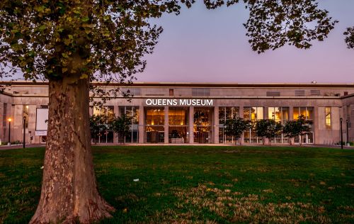 The Queens Museum building at dusk, featuring large windows and surrounded by trees and green grass.