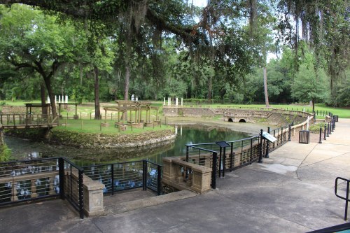 A serene park scene featuring a pond, stone pathways, and lush greenery with trees and a gazebo in the background.