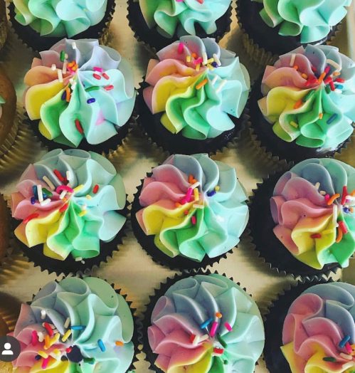 A close-up of colorful cupcakes with swirled pastel frosting and sprinkles on top, arranged in a neat pattern.