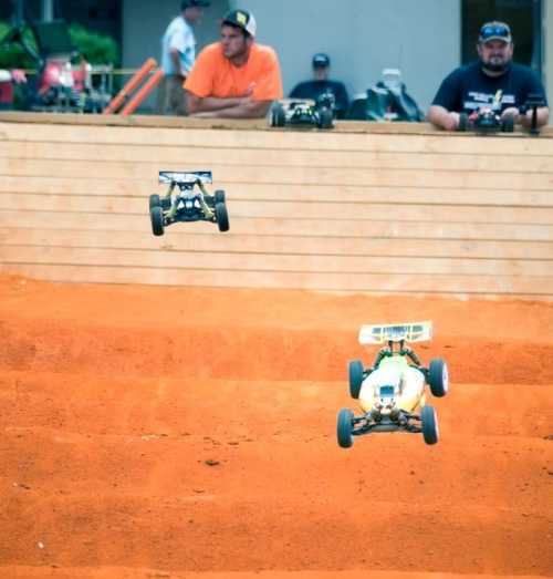 Two remote-controlled cars jumping over a dirt ramp, with spectators in the background.