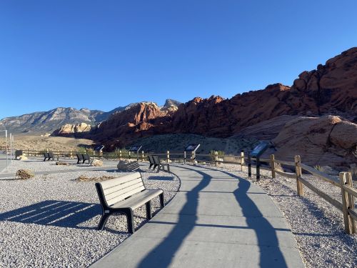A winding path with benches leads through a desert landscape, framed by colorful rock formations and clear blue skies.