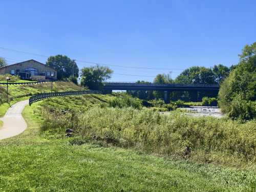 A scenic view of a riverbank with a path, bridge, and a building surrounded by greenery under a clear blue sky.