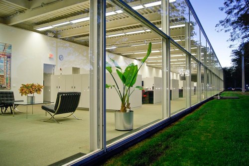 Modern office space with large windows, green plants, and minimalist furniture, set in a serene outdoor environment.
