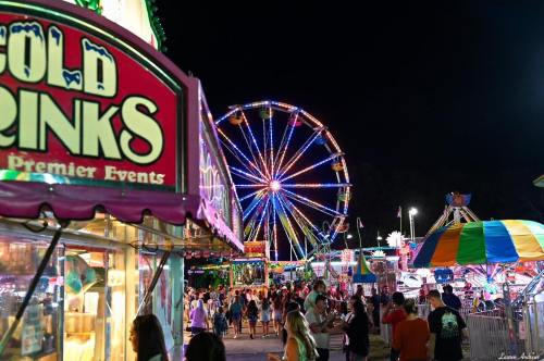 A vibrant night scene at a fair, featuring colorful lights, a Ferris wheel, and crowds enjoying various attractions.