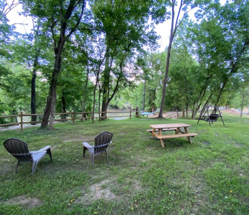 A serene outdoor scene with two chairs, a picnic table, and a swing, surrounded by trees near a river.