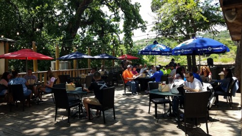 Outdoor dining area with tables under umbrellas, people enjoying meals, surrounded by trees and a sunny atmosphere.