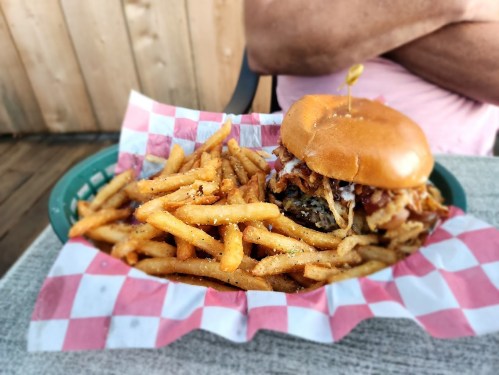 A burger topped with pulled pork and coleslaw, served with a side of golden fries in a checkered basket.