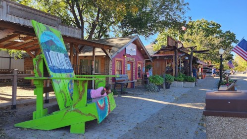 Colorful storefronts line a sunny street, featuring a large green chair and American flags in a charming outdoor setting.