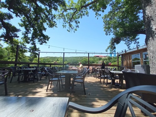 Outdoor dining area with wooden deck, tables, and chairs, surrounded by trees and a clear blue sky.