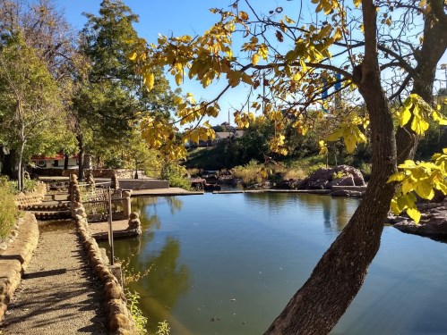 A serene pond surrounded by trees with autumn leaves, featuring a stone path and a clear blue sky.