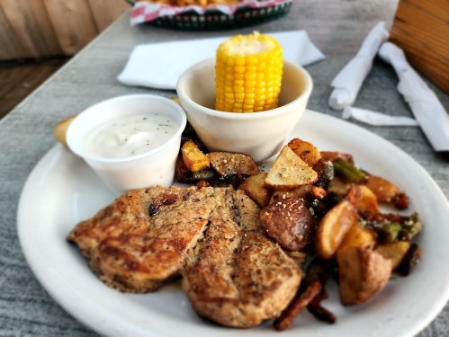 A plate of grilled pork chops, seasoned potatoes, corn on the cob, and a side of creamy dipping sauce.