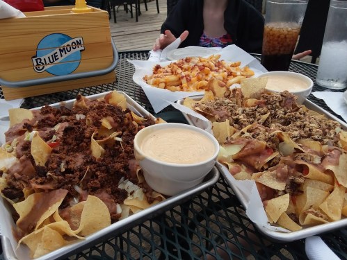 Two large trays of nachos topped with meat and cheese, alongside a plate of loaded fries, on a table at a restaurant.