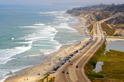 Aerial view of a coastal road alongside a beach with waves, cars parked, and people enjoying the shoreline.