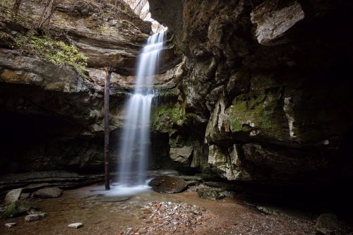 A serene waterfall cascades over rocky cliffs into a tranquil pool, surrounded by lush greenery and natural stone formations.