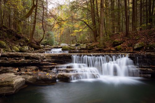 A serene waterfall cascades over rocky ledges, surrounded by autumn foliage and lush green trees in a tranquil forest setting.