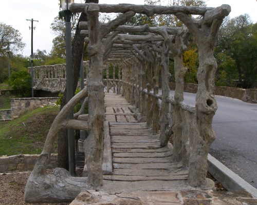 A rustic wooden bridge with a trellis-like structure, surrounded by greenery and a road in the background.