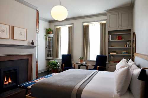 Cozy bedroom with a fireplace, large windows, and modern decor, featuring a bed, chairs, and shelves with books.