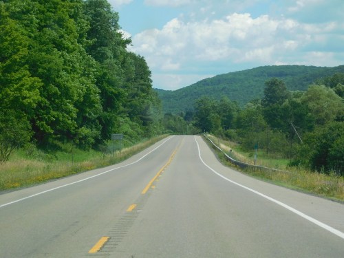 A winding road stretches through lush green trees and rolling hills under a partly cloudy sky.