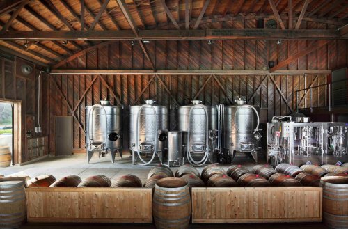Interior of a rustic winery featuring stainless steel fermentation tanks and wooden barrels in a spacious barn setting.