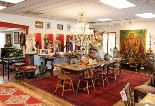 A vintage shop interior featuring a large dining table, ornate chandelier, eclectic decor, and various artworks on display.