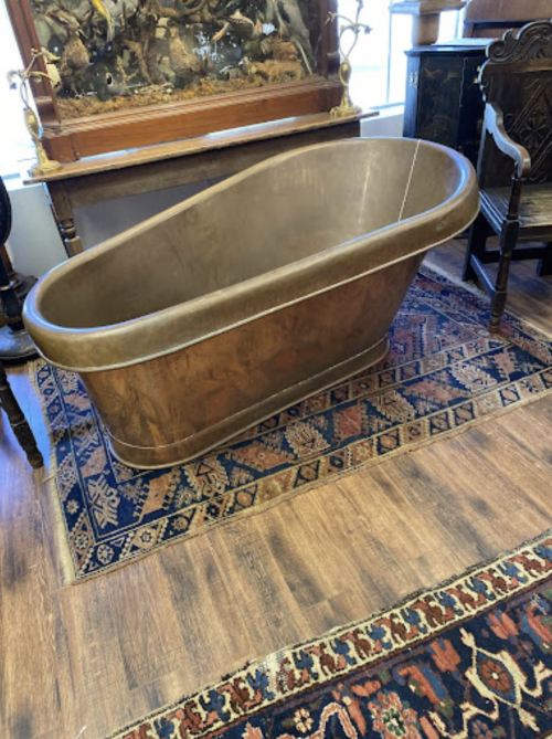 A vintage, oval-shaped metal bathtub sits on a patterned rug in a room with antique furniture.