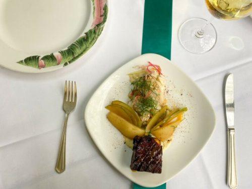 A plate of grilled meat with pickled vegetables and sliced apples, accompanied by a fork and a glass of wine.