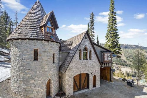 A whimsical stone castle with a turret, surrounded by trees and mountains under a clear blue sky.