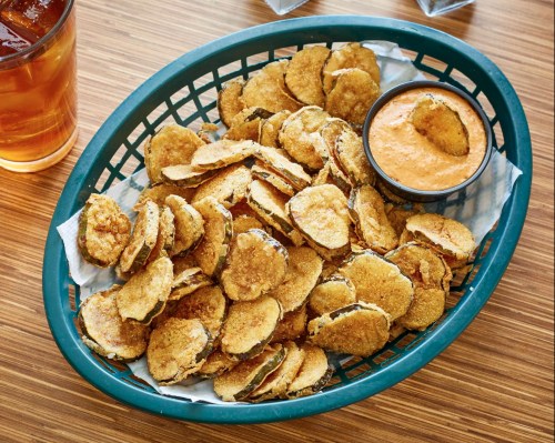 A green basket filled with crispy fried pickles and a small bowl of dipping sauce, alongside a glass of iced tea.