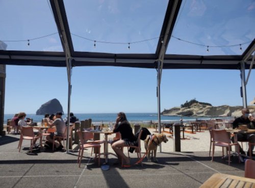A beachside restaurant with diners, a dog, and a view of the ocean and rocky cliffs under a clear blue sky.