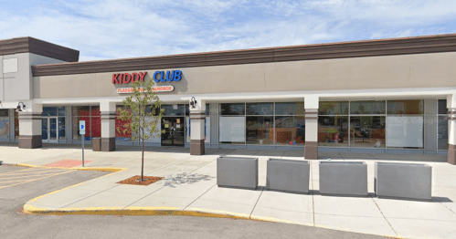 Exterior of a storefront labeled "Kiddy Club," featuring large windows and a sidewalk with planters.