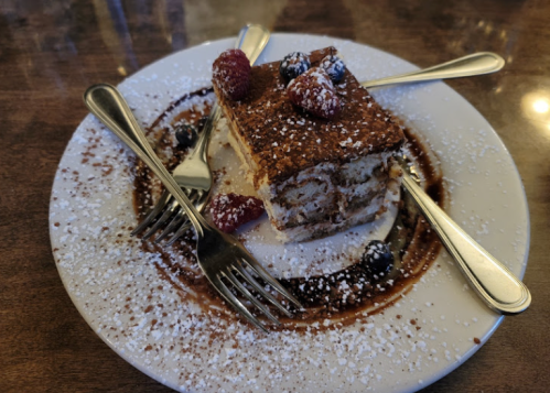 A slice of tiramisu on a plate, garnished with raspberries and blueberries, surrounded by powdered sugar and chocolate sauce.