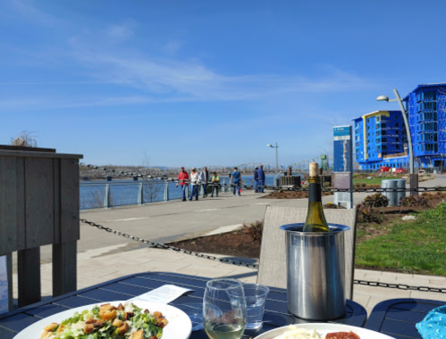 A table with food and drinks overlooks a waterfront path where people stroll, with blue buildings in the background.