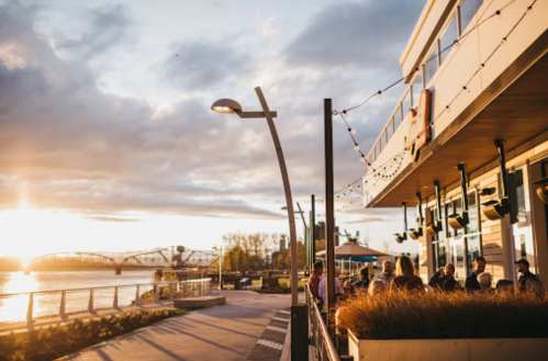 A riverside restaurant at sunset, with outdoor seating and decorative lights, creating a warm, inviting atmosphere.