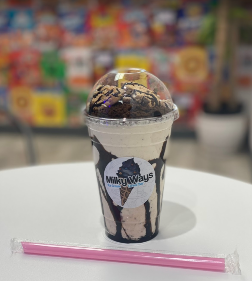 A large milkshake topped with whipped cream and chocolate drizzle, served in a clear cup with a pink straw.
