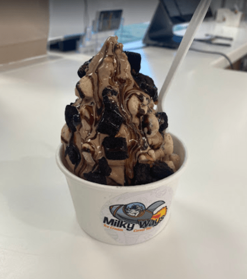 A cup of chocolate soft serve topped with crushed Oreos and chocolate drizzle, served with a white spoon.