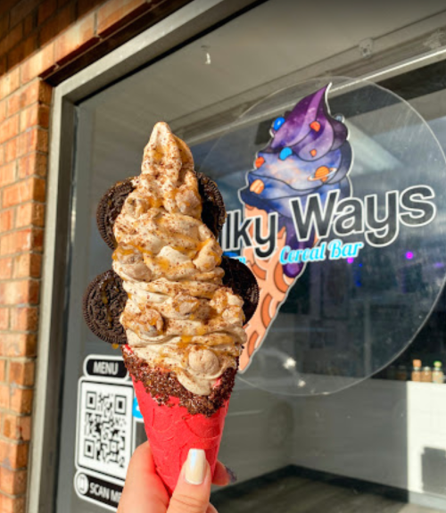 A hand holding a swirl ice cream cone topped with cookies, in front of a shop window with a colorful ice cream logo.
