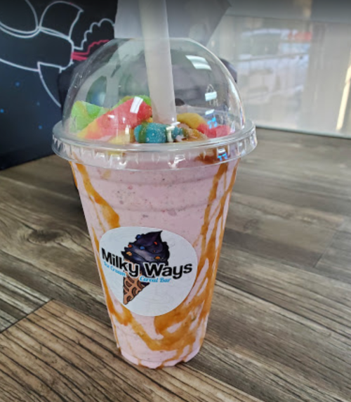 A colorful milkshake topped with candy, served in a clear cup with a logo for Milky Ways Dessert Bar.