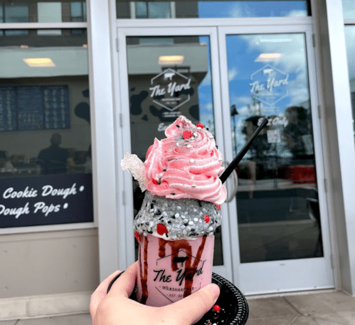 A hand holds a colorful milkshake topped with pink and gray whipped cream, in front of a shop entrance.